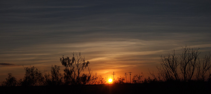 [In the foreground are open bushes, in the midground are power poles, and in the distance is the sun just at the horizon. The thin layers of clouds creates a haze across the color so the sun's edges are blurred. There is a section of orange clouds around the sun and white and grey hazes of clouds above it.]